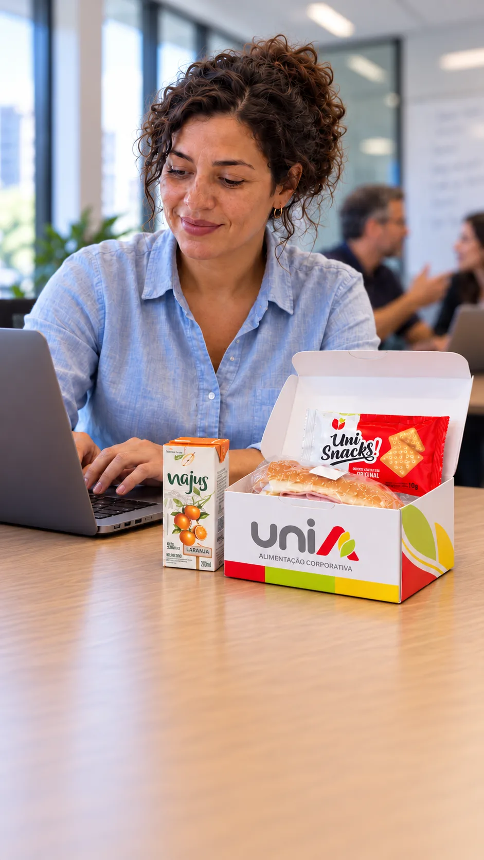 Profissional trabalhando no escritório com kit lanche UniA: sanduíche, Uni Snacks e suco Najus de laranja